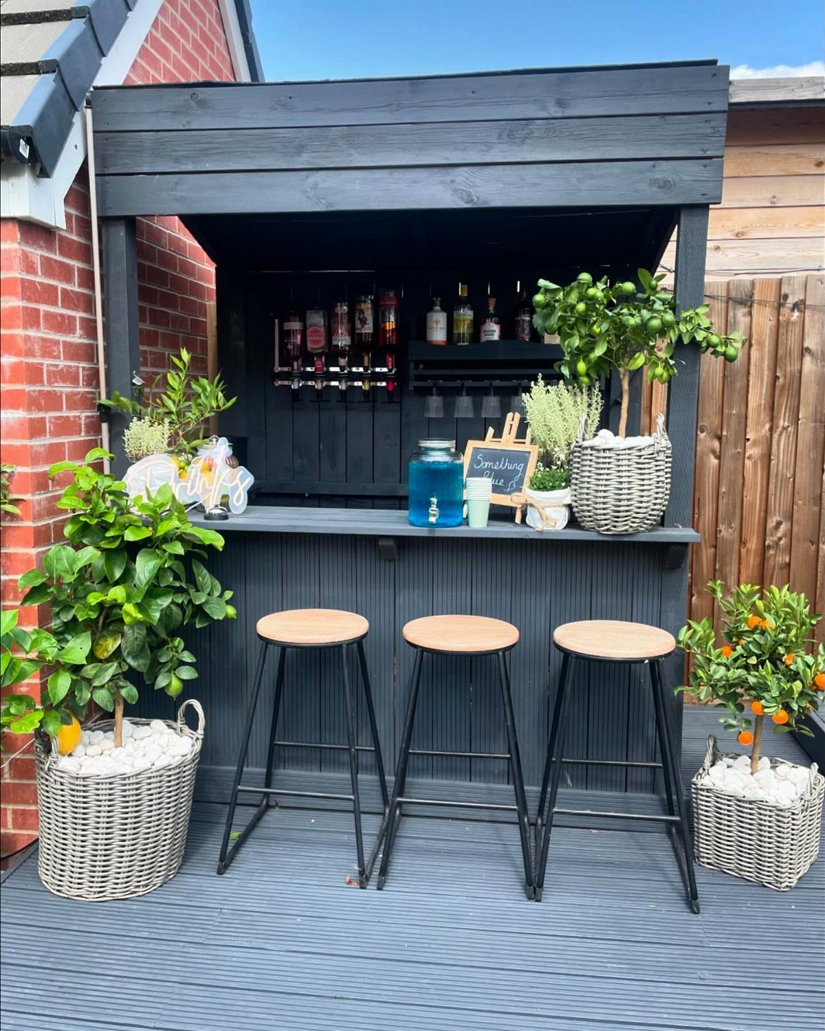 23 Backyard DIY Ideas for a Patio, Garden and Fire Pit 9 Dark grey wooden outdoor bar with three stools and potted citrus plants on a deck.