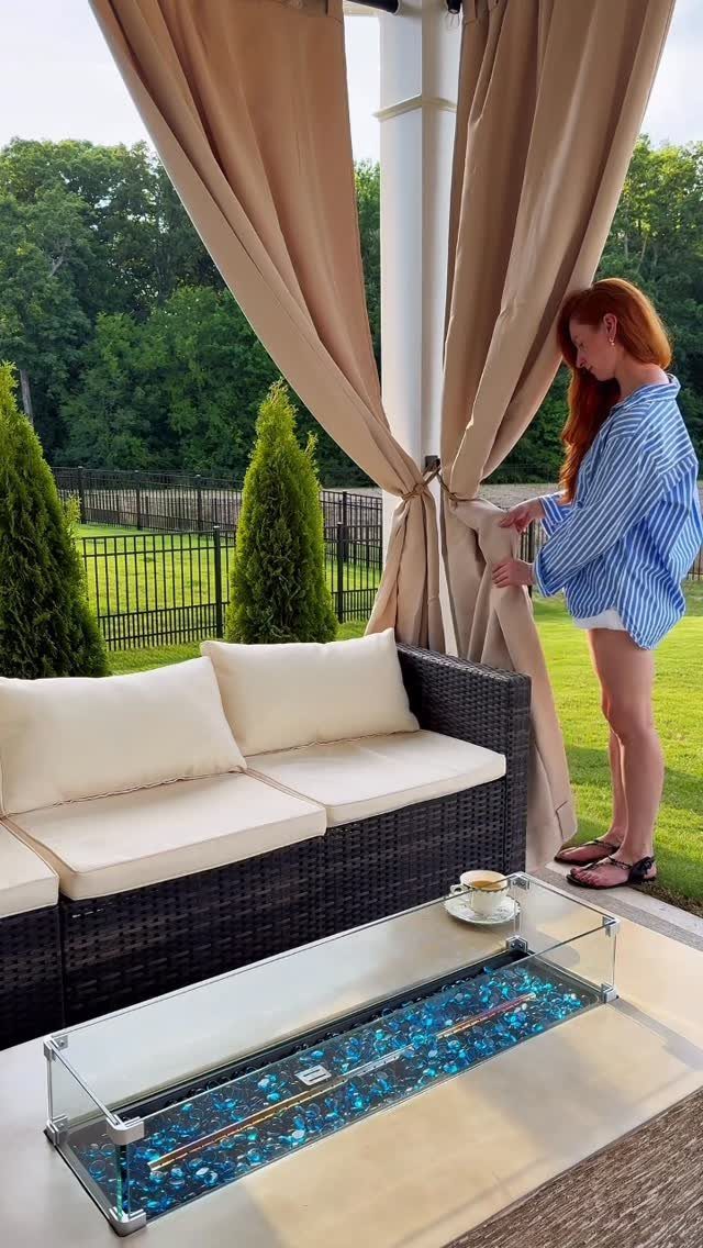 23 Backyard DIY Ideas for a Patio, Garden and Fire Pit 13 Woman adjusting tan outdoor curtains on a white pergola next to a wicker sofa.