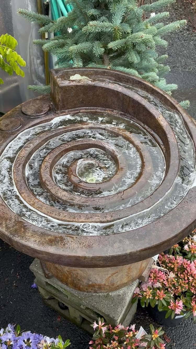 Spiral stone water fountain with flowing water surrounded by greenery and flowers.