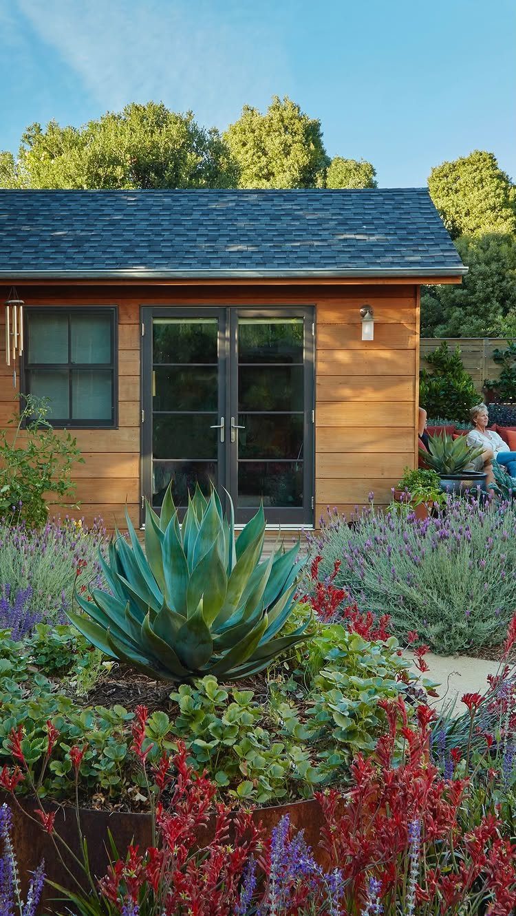 Wooden garden shed with French doors surrounded by colorful flowers and an agave plant.