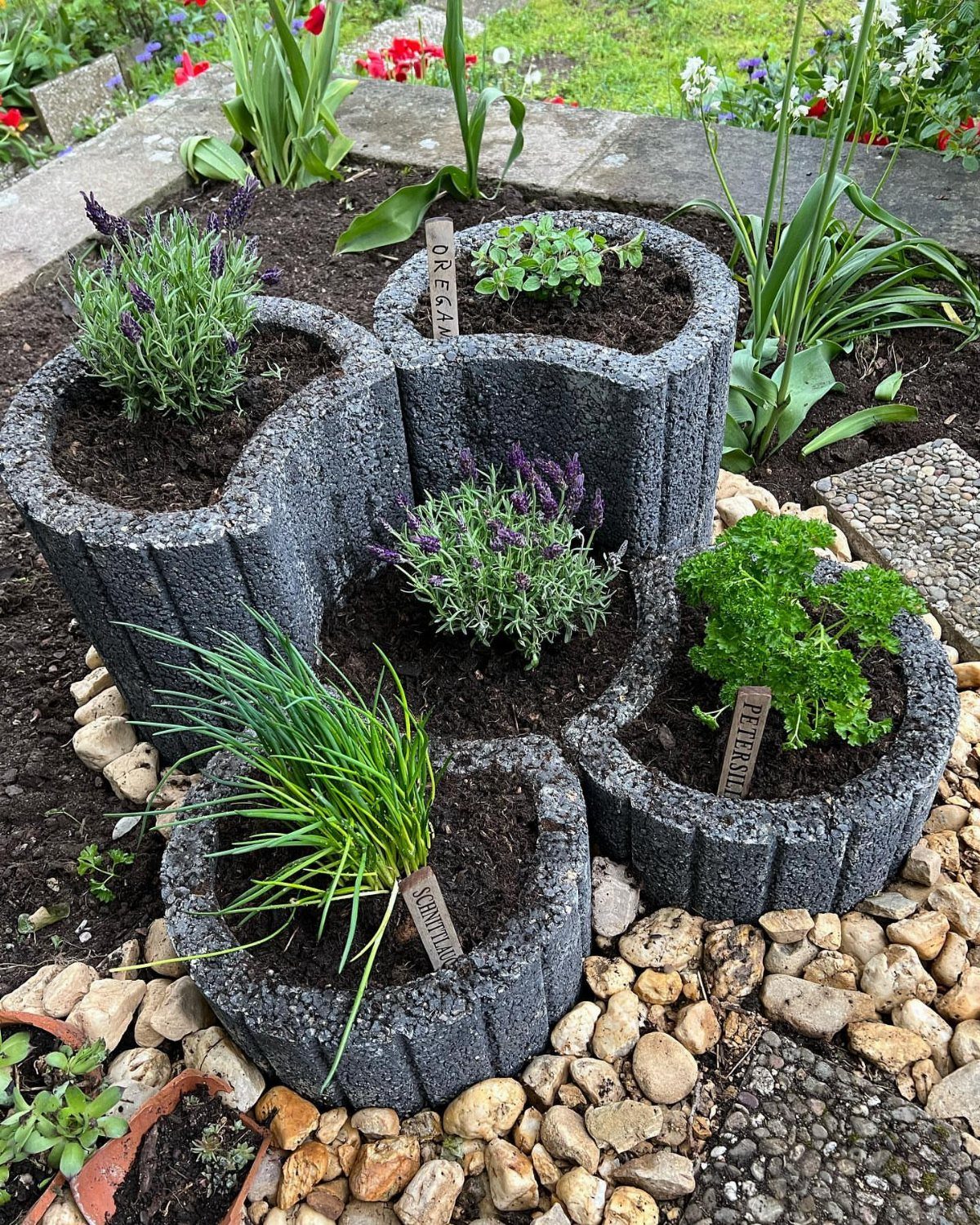 Grey concrete planters with herbs and flowers sitting on a bed of pebbles.