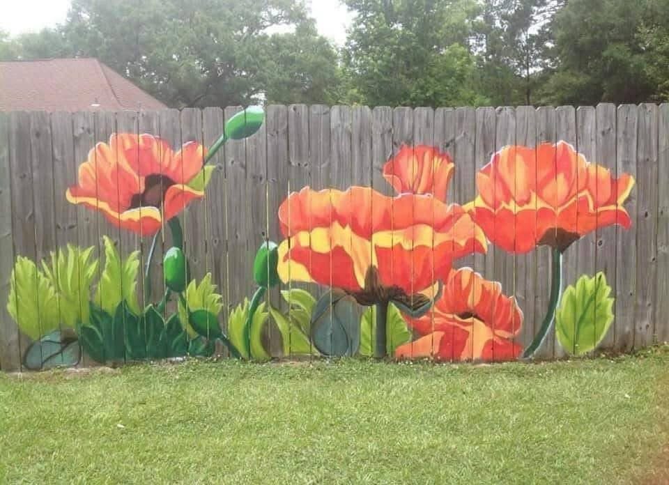 Wooden fence painted with large red and orange poppies overlooking a green lawn.