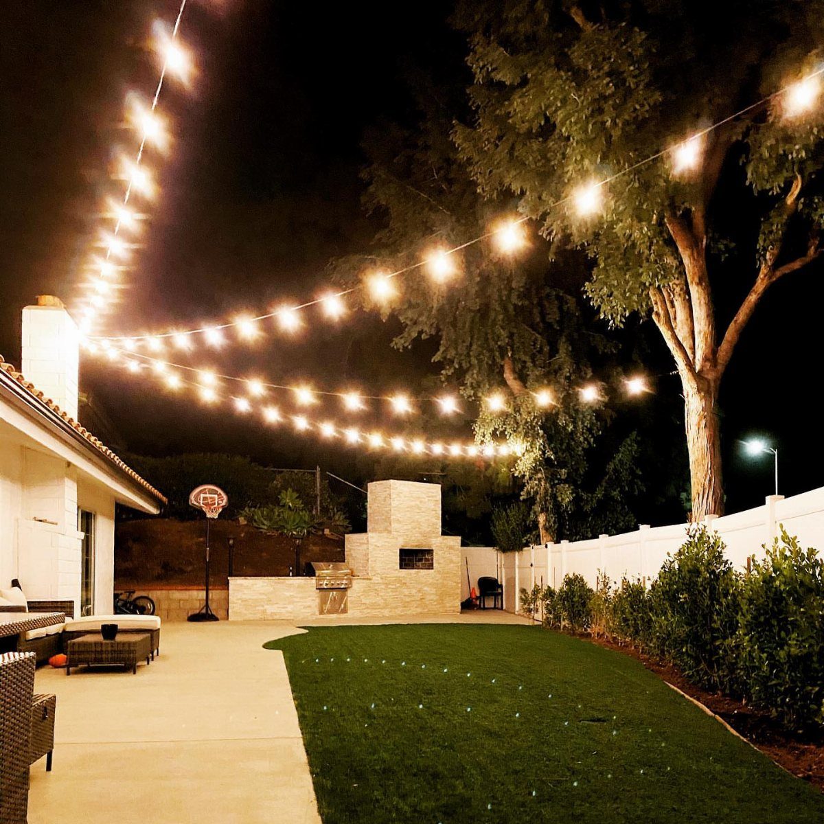 Glowing string lights crisscrossing over a backyard patio and lawn at night.