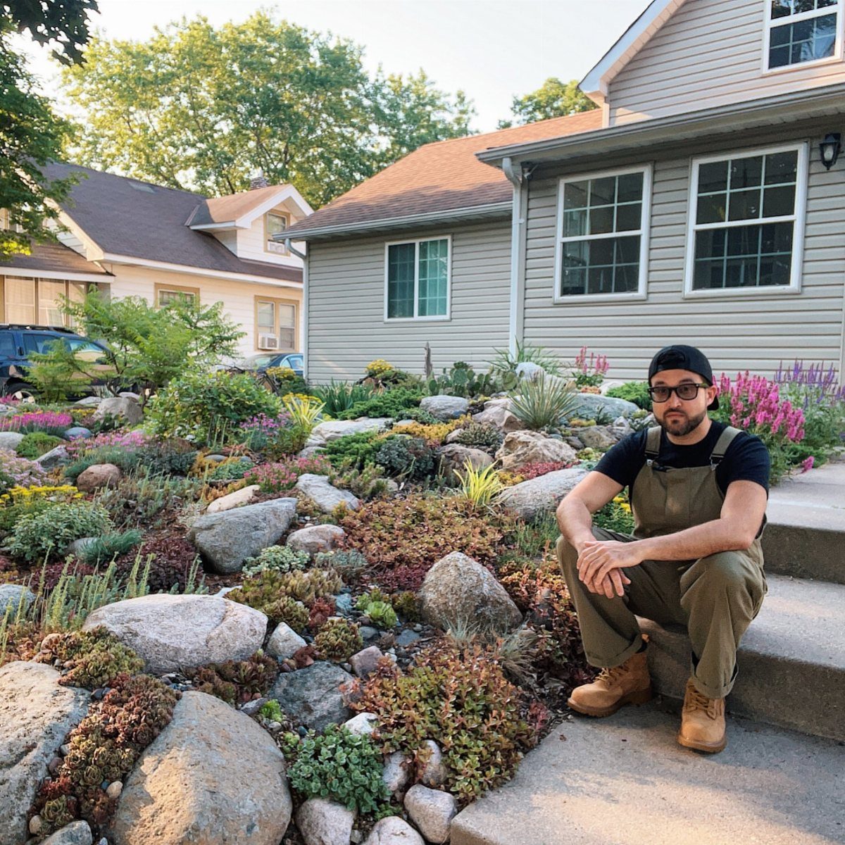 14 Cheap Garden Makeover Ideas to Refresh Your Yard 10 Man in overalls posing next to a landscaped rock garden with stones and succulents.