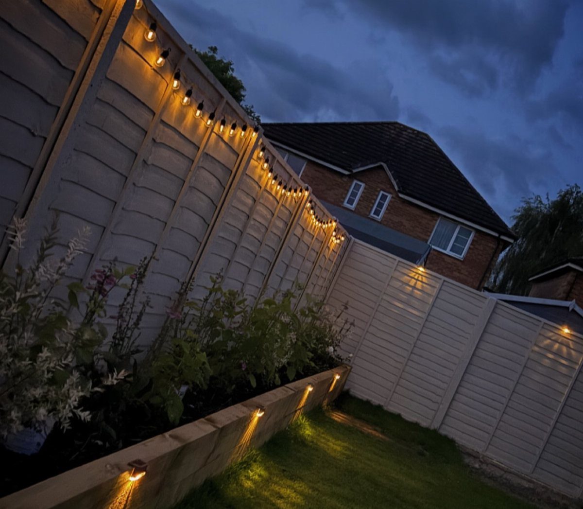 14 Cheap Garden Makeover Ideas to Refresh Your Yard 3 String lights and solar border lights illuminating a wooden fence and garden at dusk.