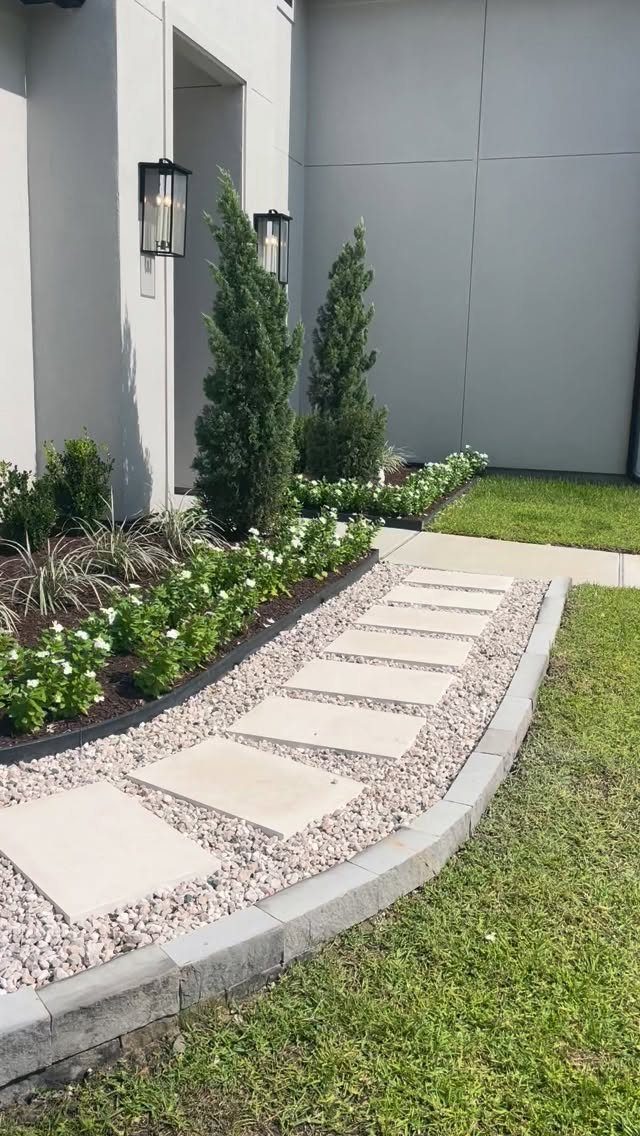 Landscaped garden bed with conical evergreens and a stone paver pathway along a house.