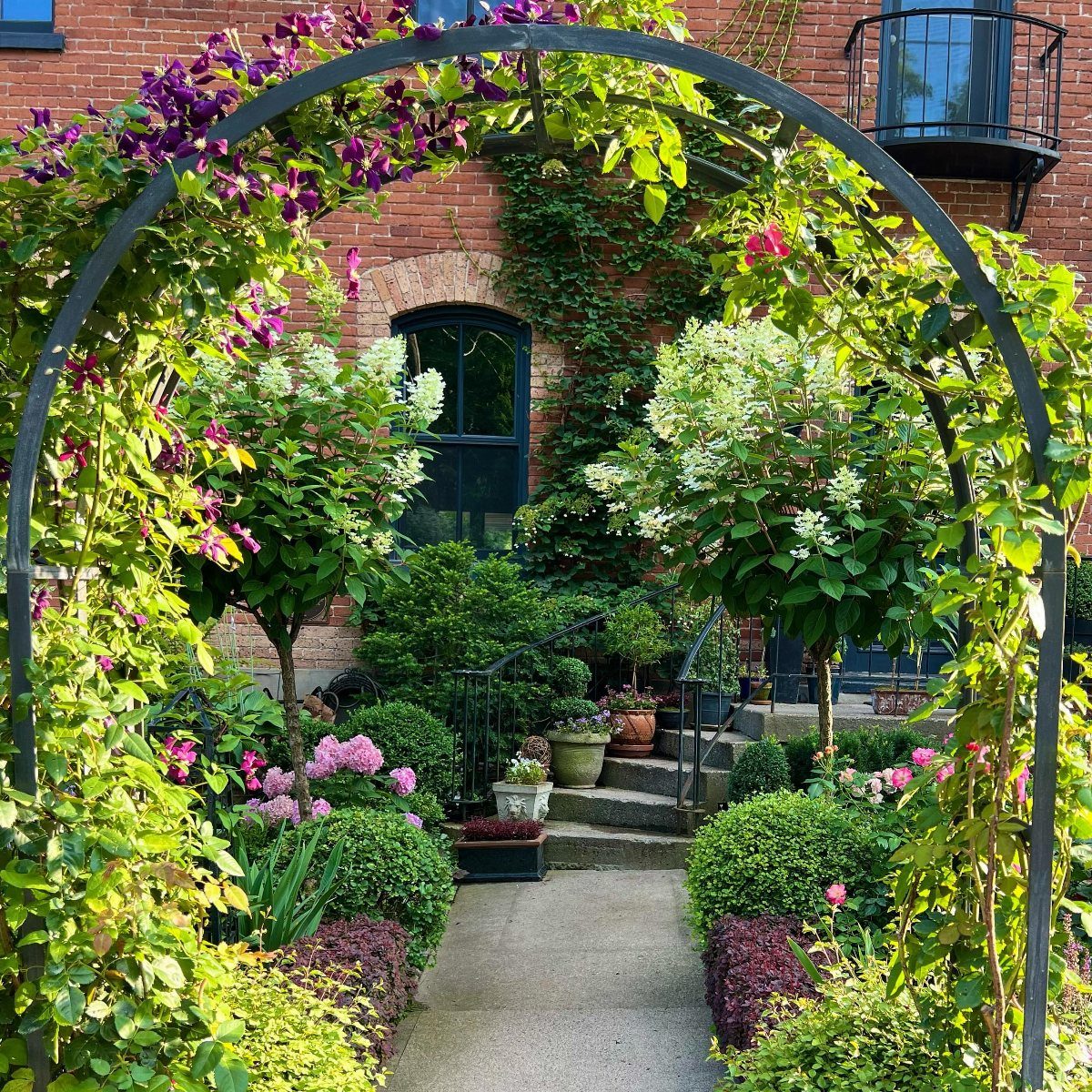 Black metal garden arbor arch framing a path with lush flowers and greenery.