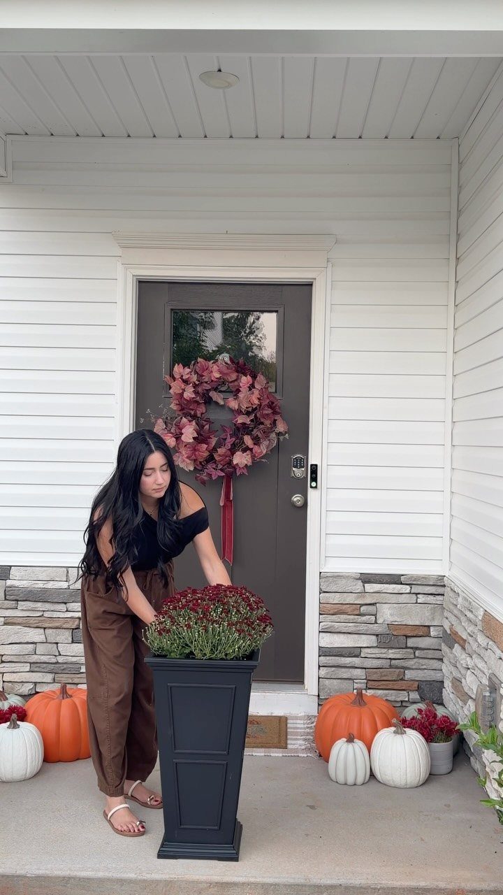 Woman arranging red mums in a tall black planter on a house porch.