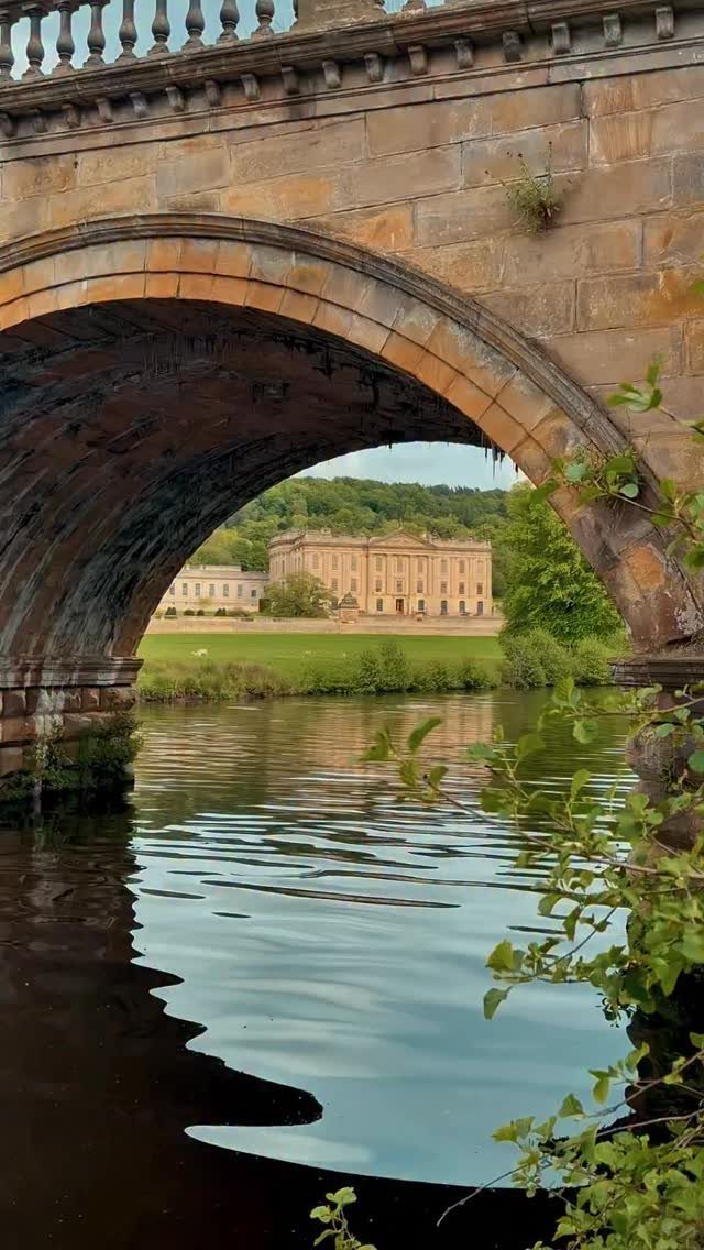 Stone manor house viewed through a bridge arch over a calm river.