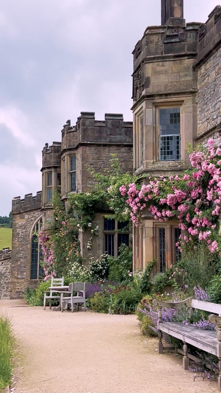Stone castle facade with pink climbing roses and wooden benches along a gravel path.