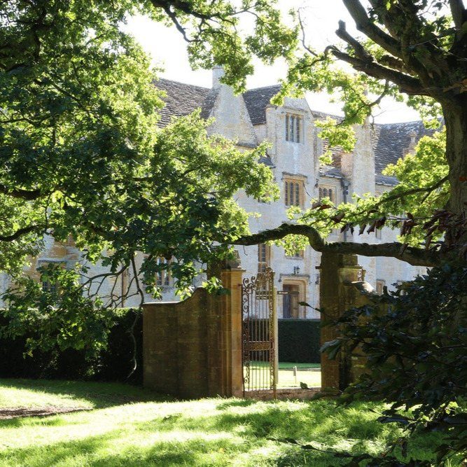 Stone manor house seen through green tree branches with an open iron gate.