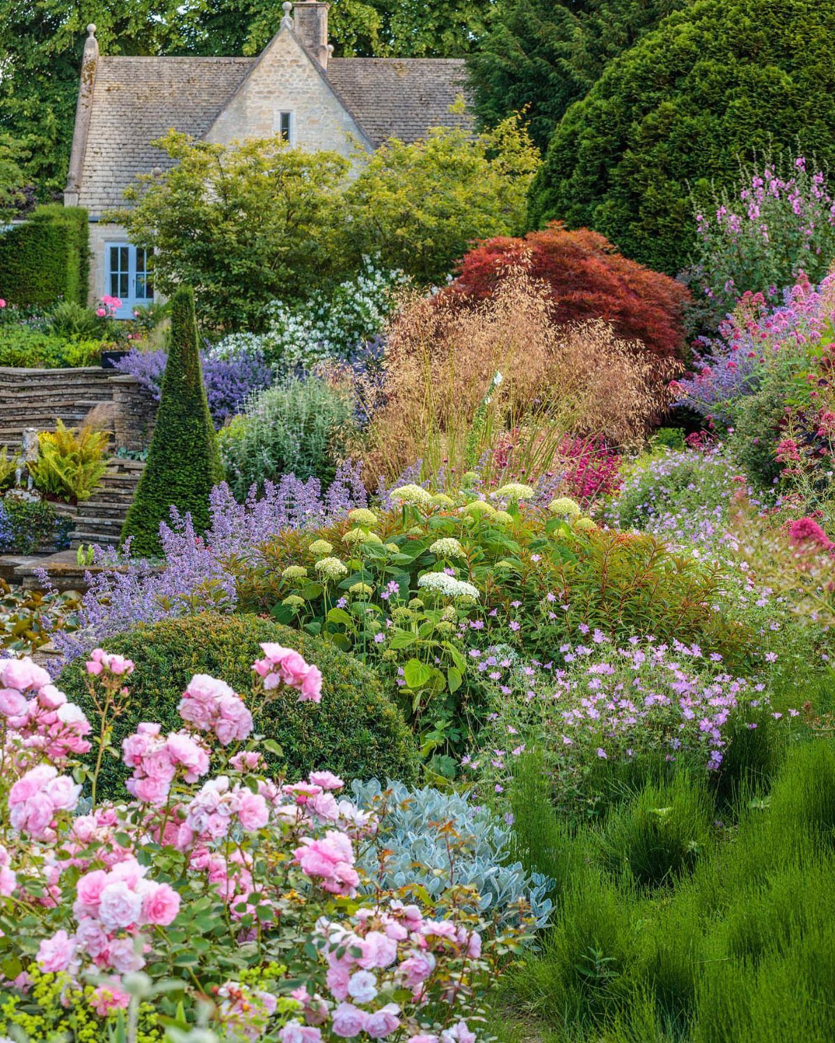 English Garden Design Ideas: 10 Classic Elements to Use 2 Curved garden beds filled with pink roses and purple flowers leading to a cottage.