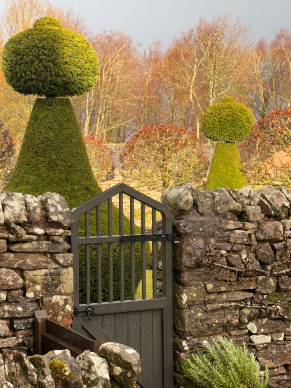 English Garden Design Ideas: 10 Classic Elements to Use 5 Weathered stone wall and wooden gate with large topiary shrubs and autumn trees.