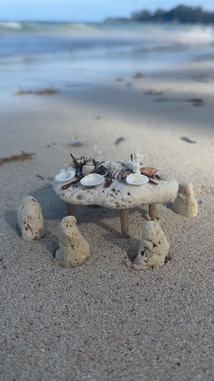 Miniature stone table with seashells on a sandy beach near the ocean waves.