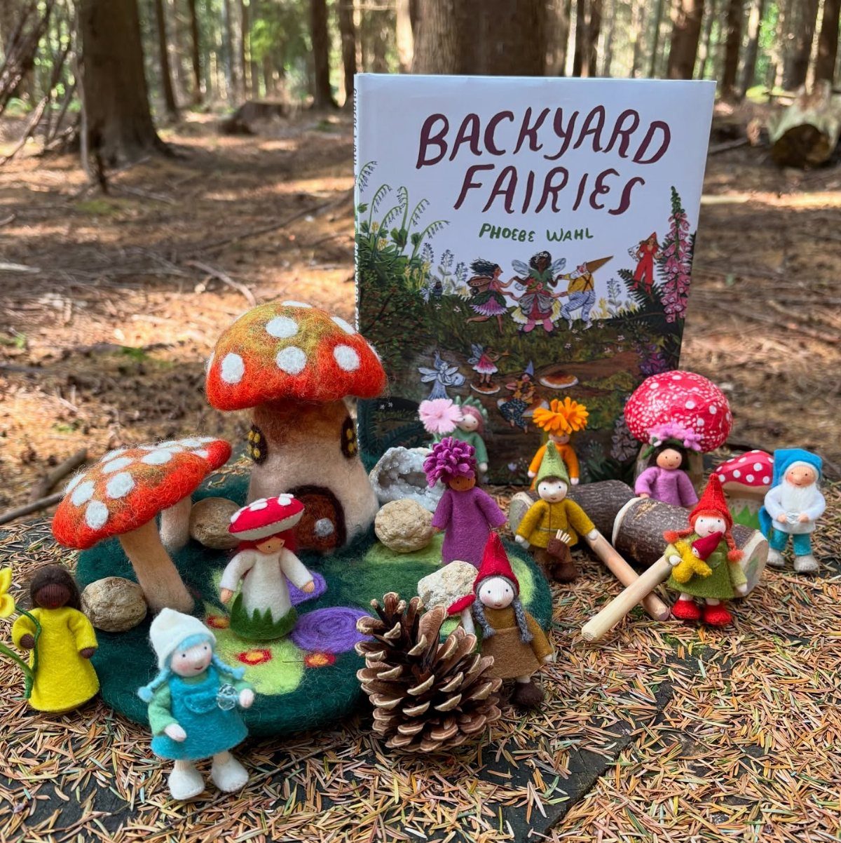 Felt fairy figurines and mushroom houses arranged on a forest floor with a book.