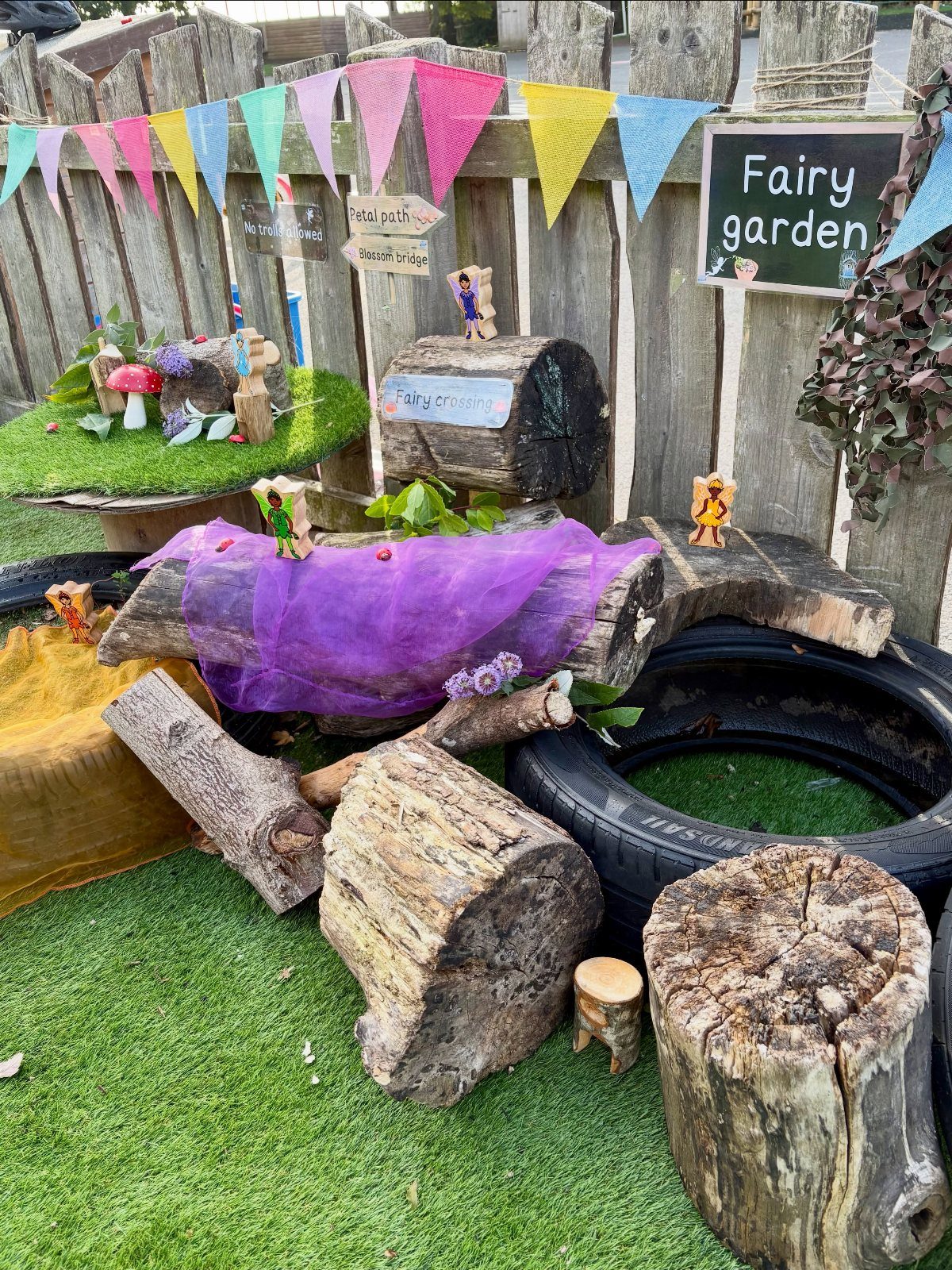 Miniature fairy garden with logs, figurines, and colorful bunting against a wooden fence.