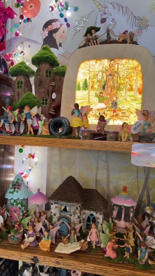 Miniature fairy garden display with small houses and winged figurines on wooden shelves.