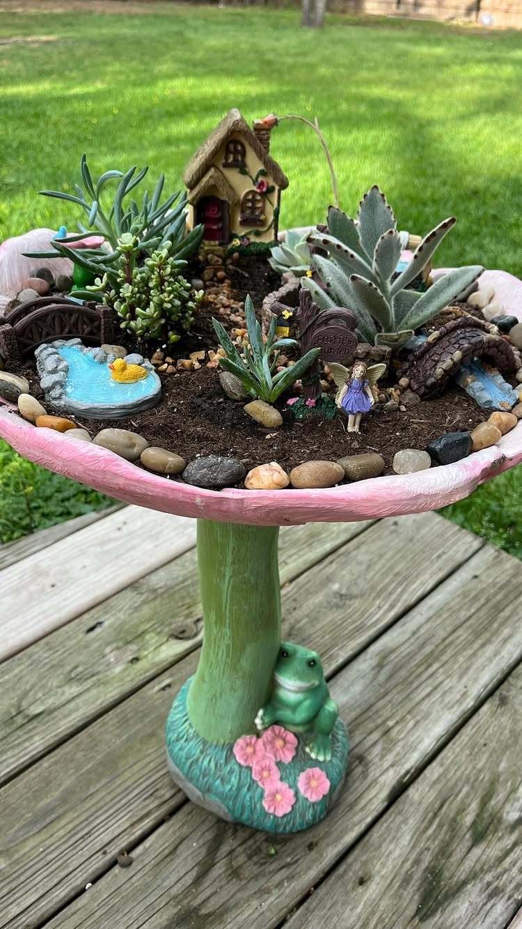 Miniature fairy garden on a pink mushroom pedestal with a small cottage and succulents.