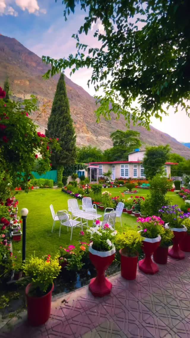 Lush green garden with white patio furniture and red flower pots near a mountain.