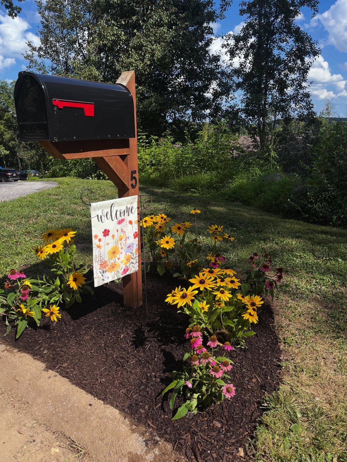 Black mailbox on a wooden post surrounded by yellow and pink flowers in mulch.