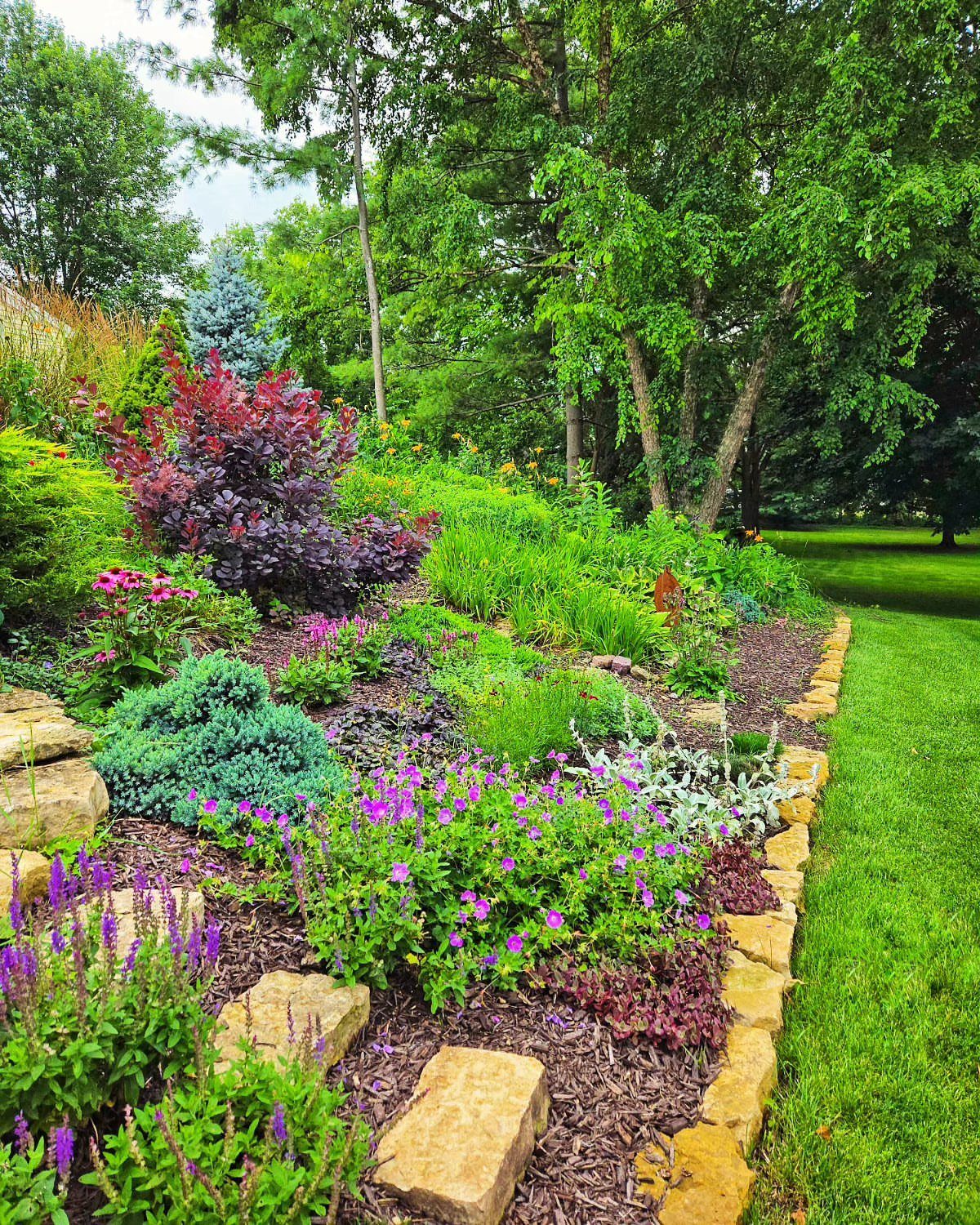 Sloped flower bed with colorful perennials and stone edging next to a green lawn.