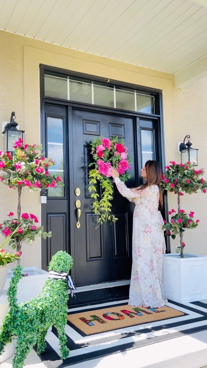 26 Front Garden Ideas for Curb Appeal and Small Yards 12 Woman arranging a pink floral wreath on a black front door with garden decor.