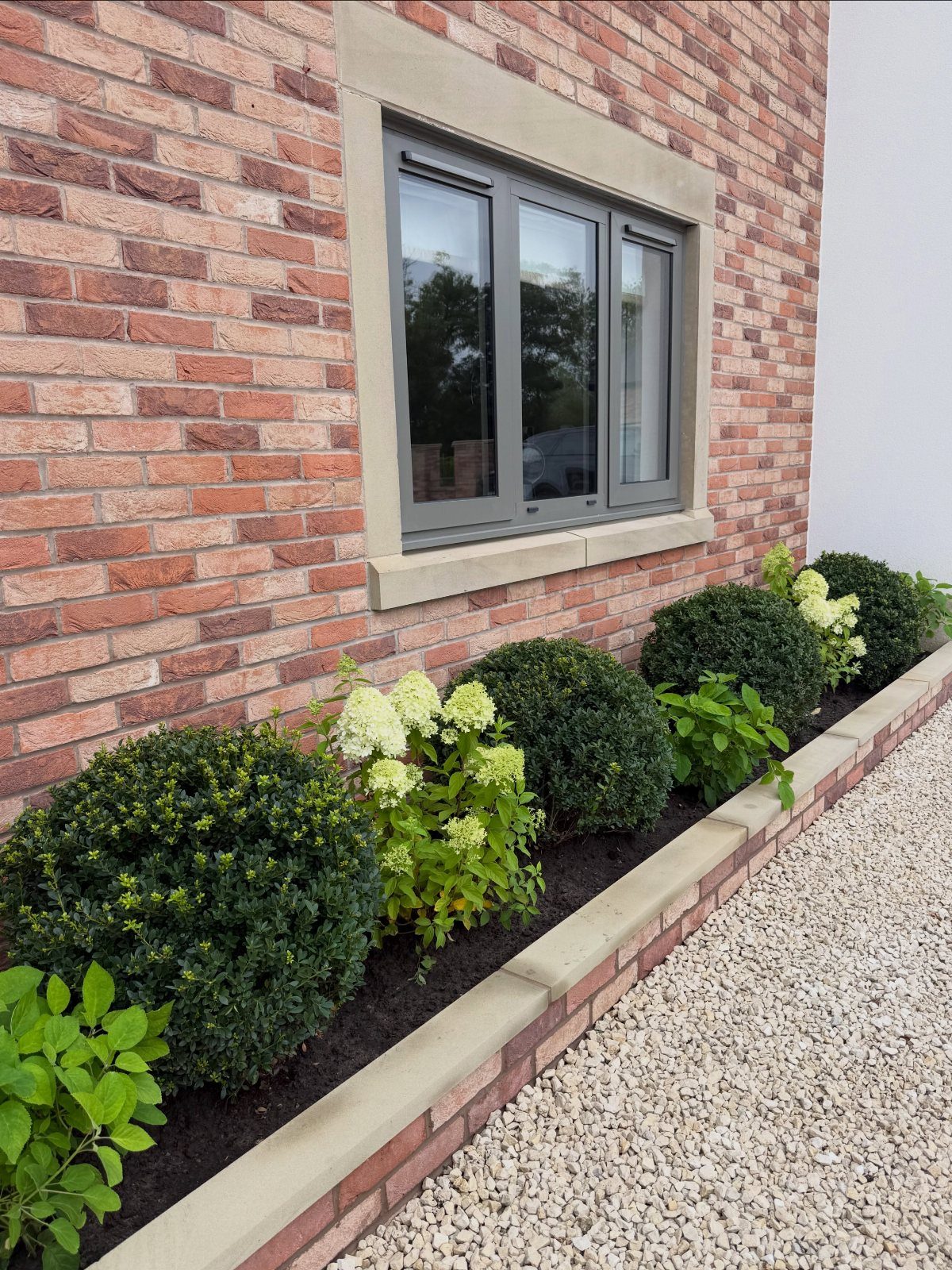 26 Front Garden Ideas for Curb Appeal and Small Yards 19 Raised brick garden bed with green shrubs and white hydrangeas along a gravel path.