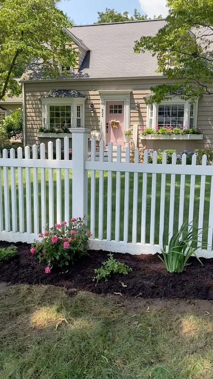 White picket fence with a dark mulch flower bed and pink blossoms in front.
