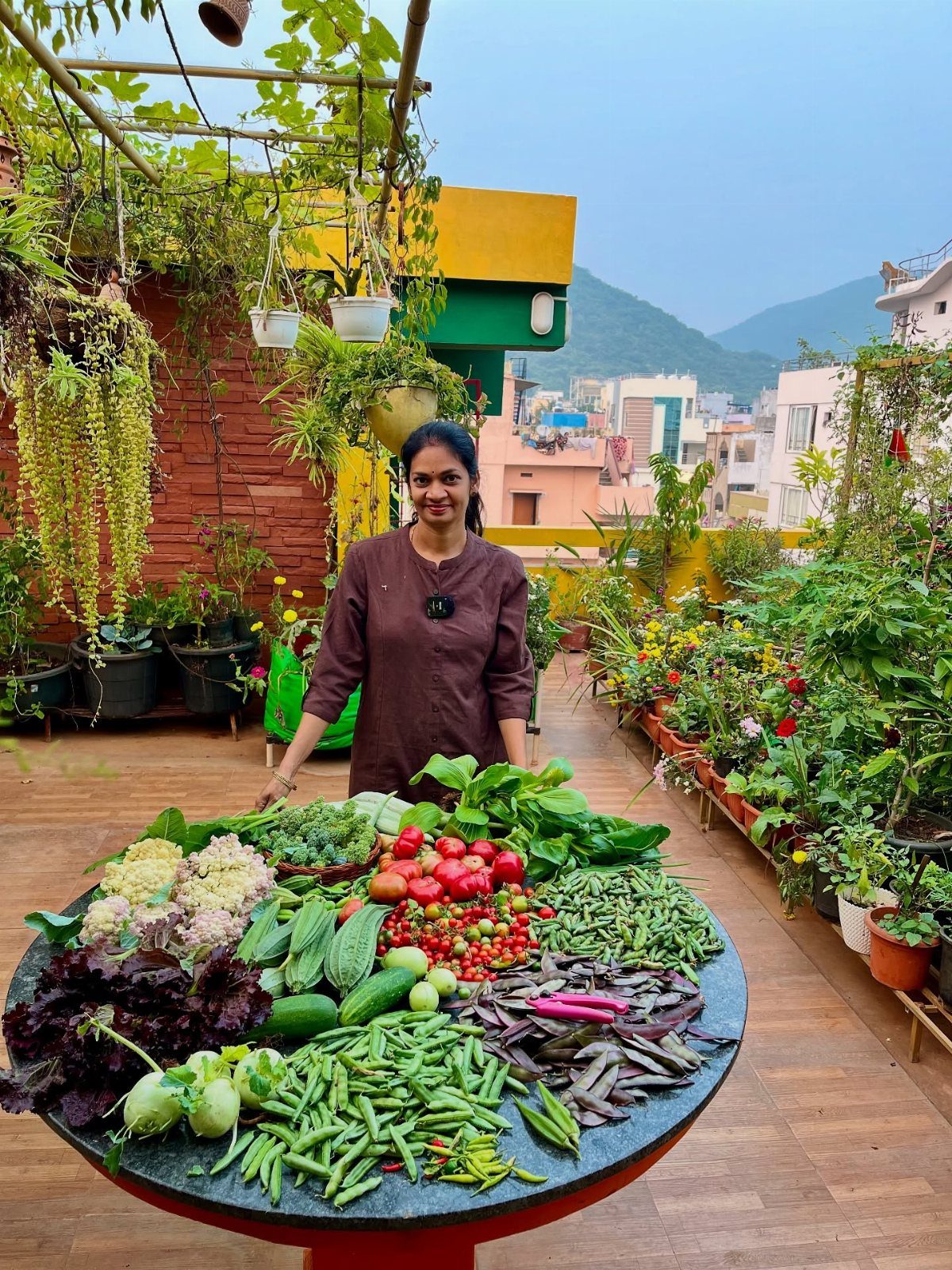 Woman standing behind a table of harvested vegetables on a lush city rooftop garden.