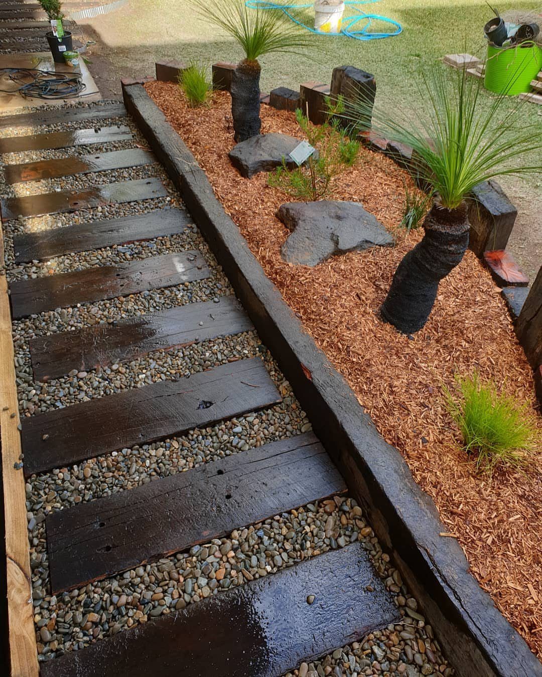 Railway sleeper path with grey pebbles and a mulched garden bed with grass trees.