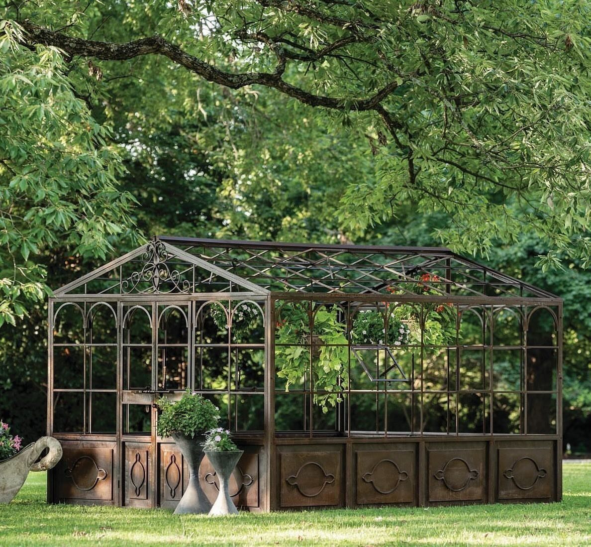 Ornate metal greenhouse with glass panels standing on a green lawn under trees.