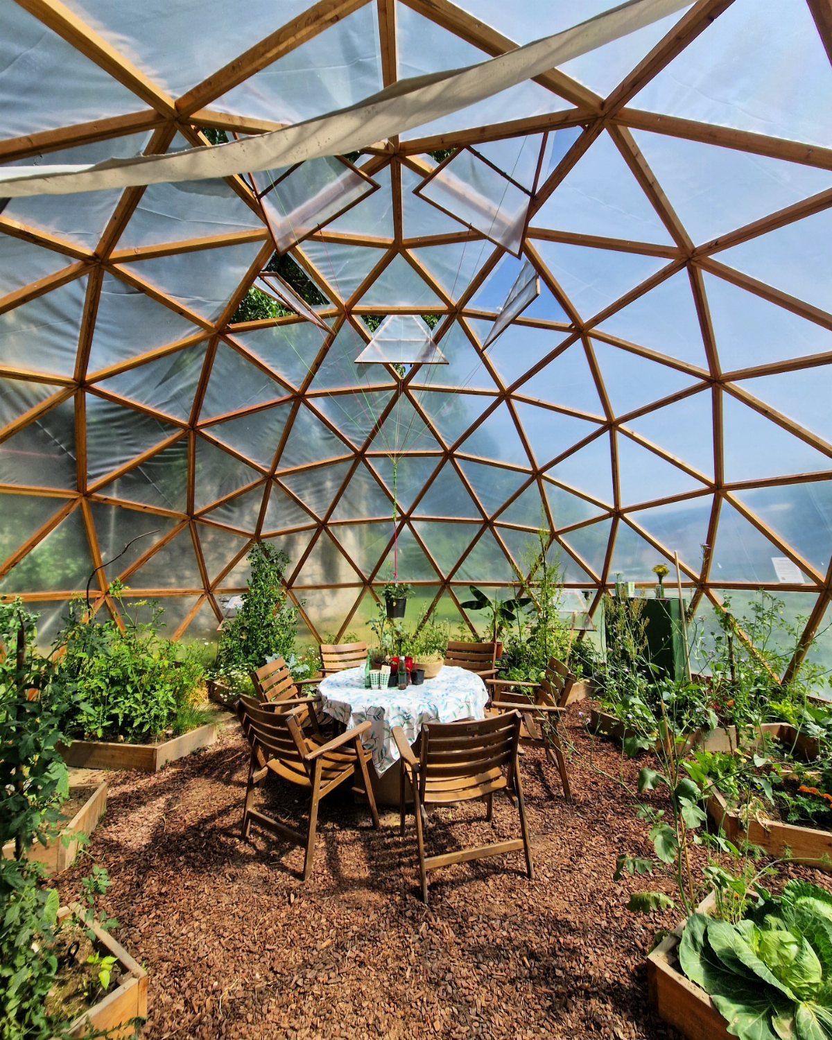 Geodesic dome greenhouse interior with wooden framing, garden beds, and a small dining set.
