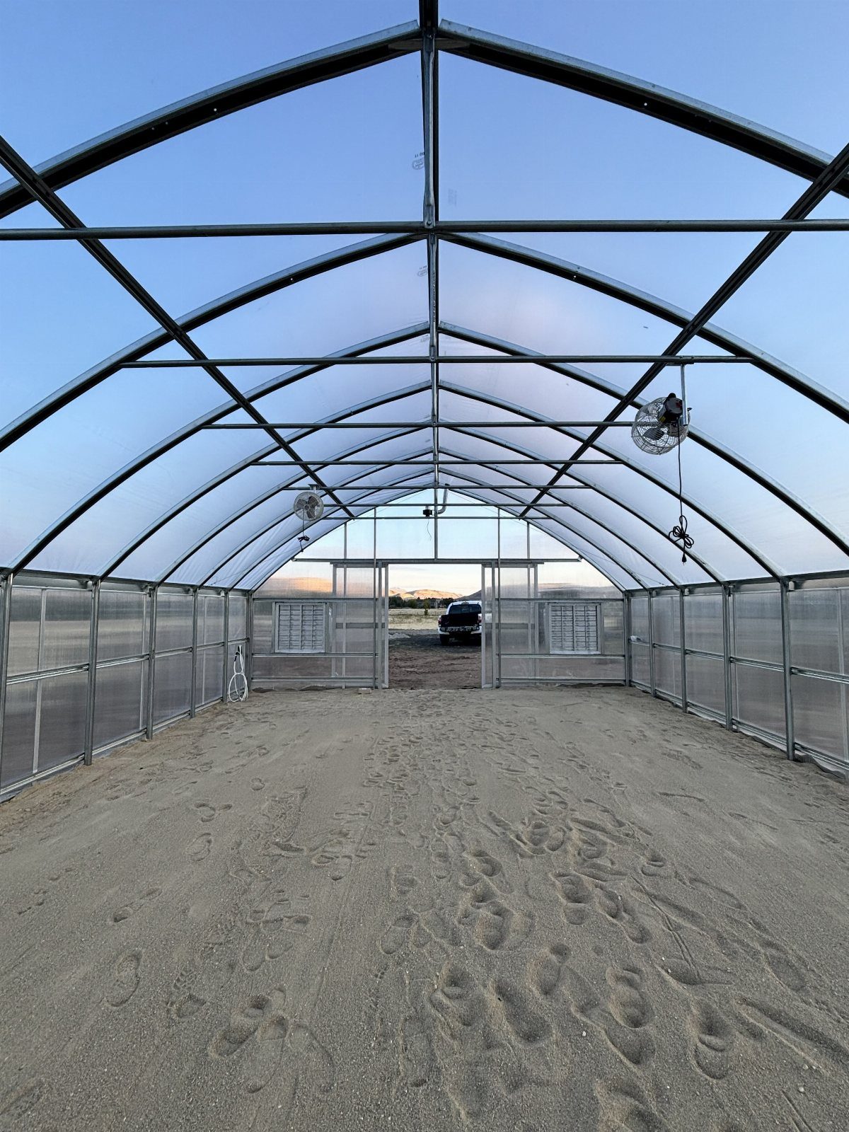 Gothic arch greenhouse interior with metal framing and sandy dirt floor.