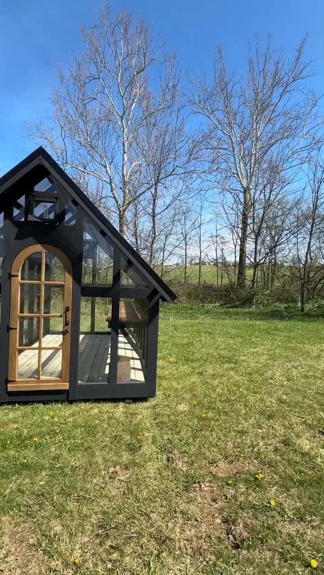 Modern black glass greenhouse with an arched wooden door on a grassy field.