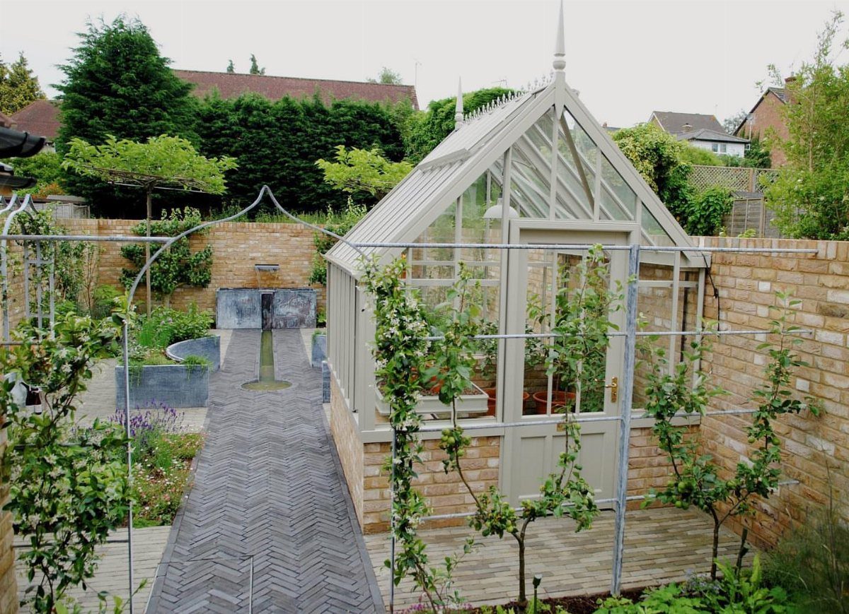 Grey herringbone paver path leading toward a small glass greenhouse and brick walls.
