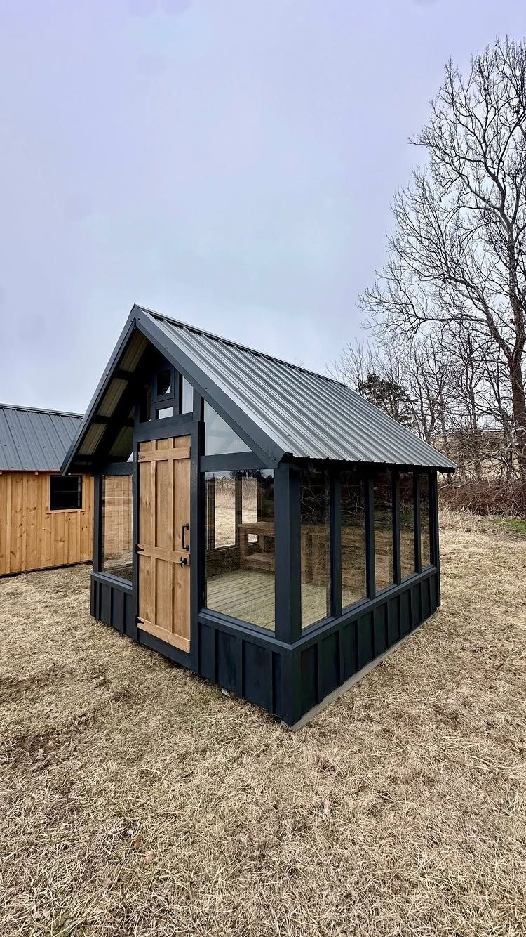 Small greenhouse with black walls, glass panels, and a natural wood door.