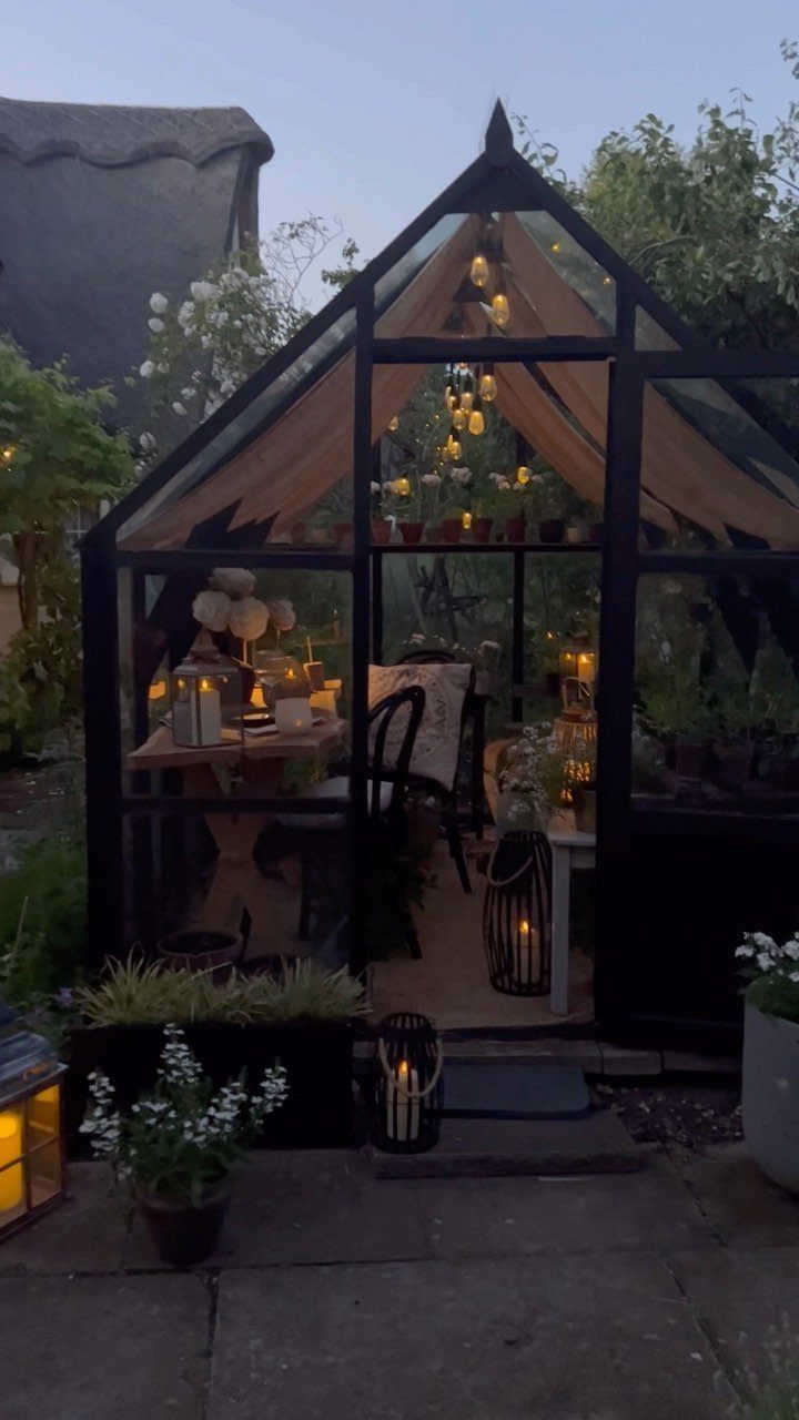 Black-framed glass cottage style greenhouse with interior lighting and garden furniture inside.