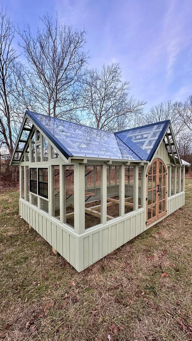 Light green wooden greenhouse with a gabled roof and arched door in a field.
