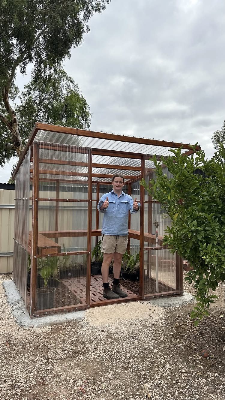 Man standing in a custom wooden greenhouse with translucent walls and a flat roof.