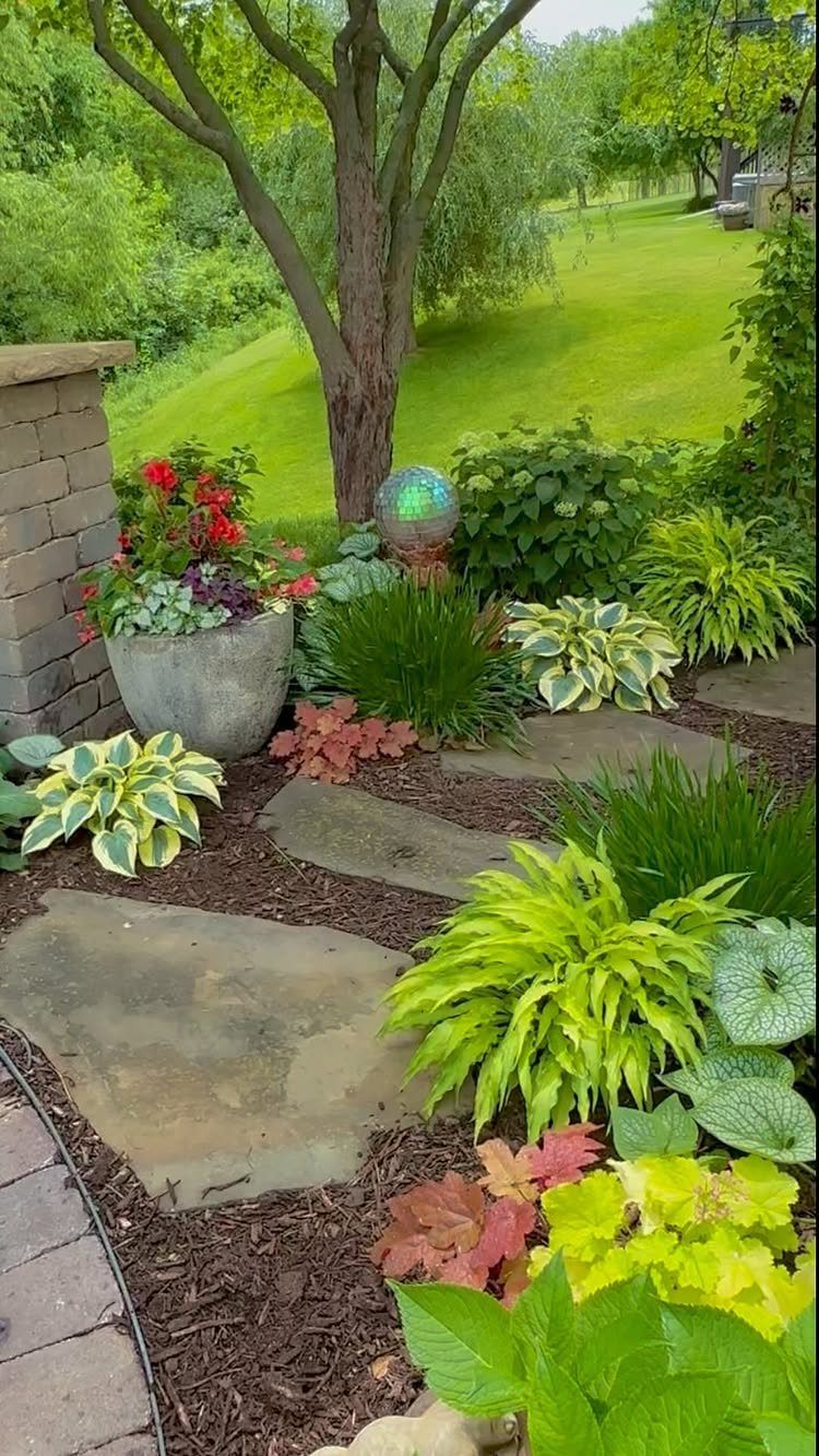 10 Ideas for Landscaping Around Trees, From Moss to Lighting 4 Landscaped garden with hostas, red flowers, and stone pavers around a tree trunk.