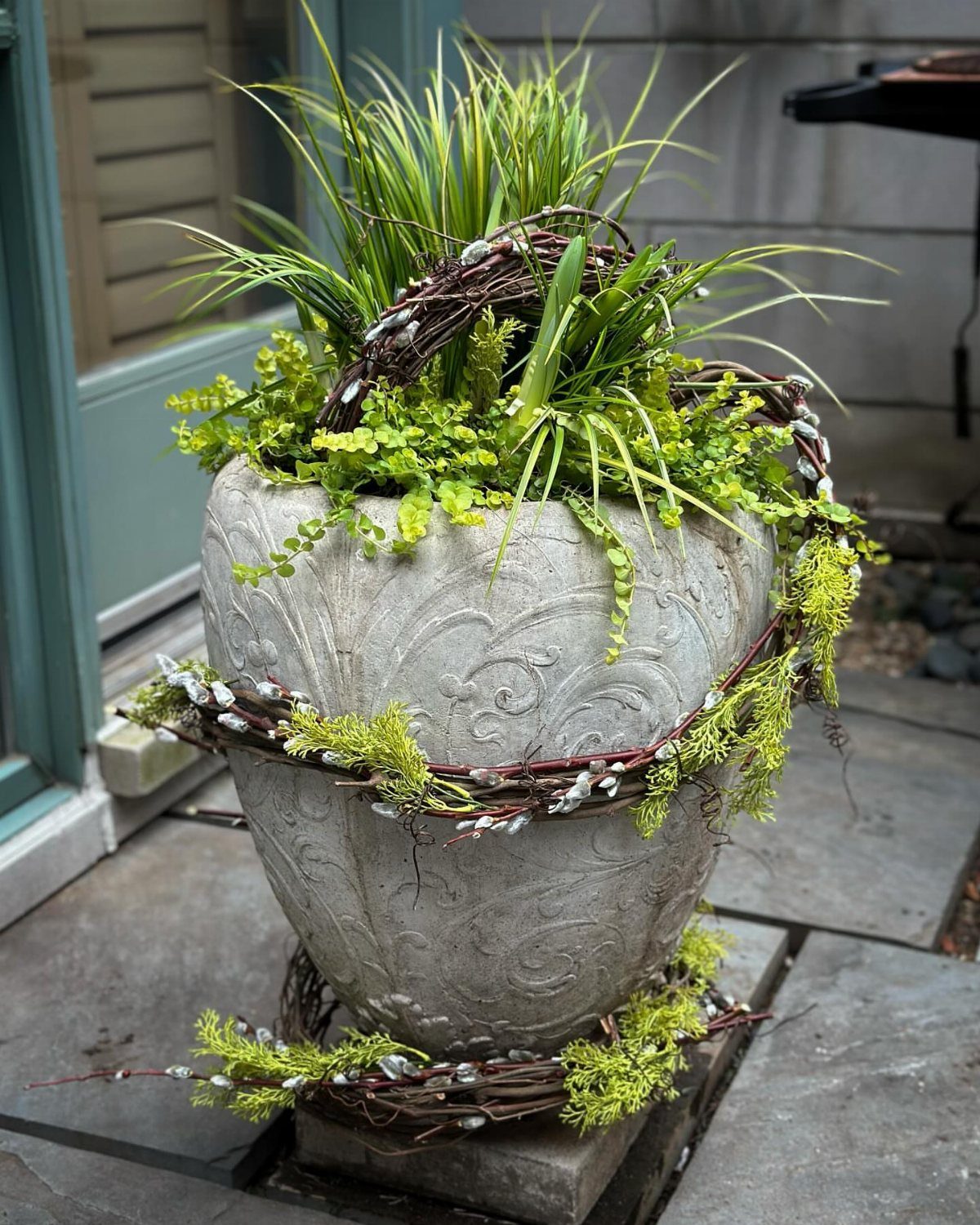Ornate grey stone planter with tall grasses and trailing green foliage on a patio.