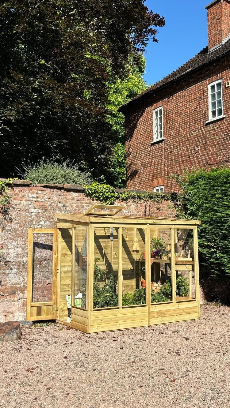 Wooden garden greenhouse with glass panels containing plants next to a brick wall