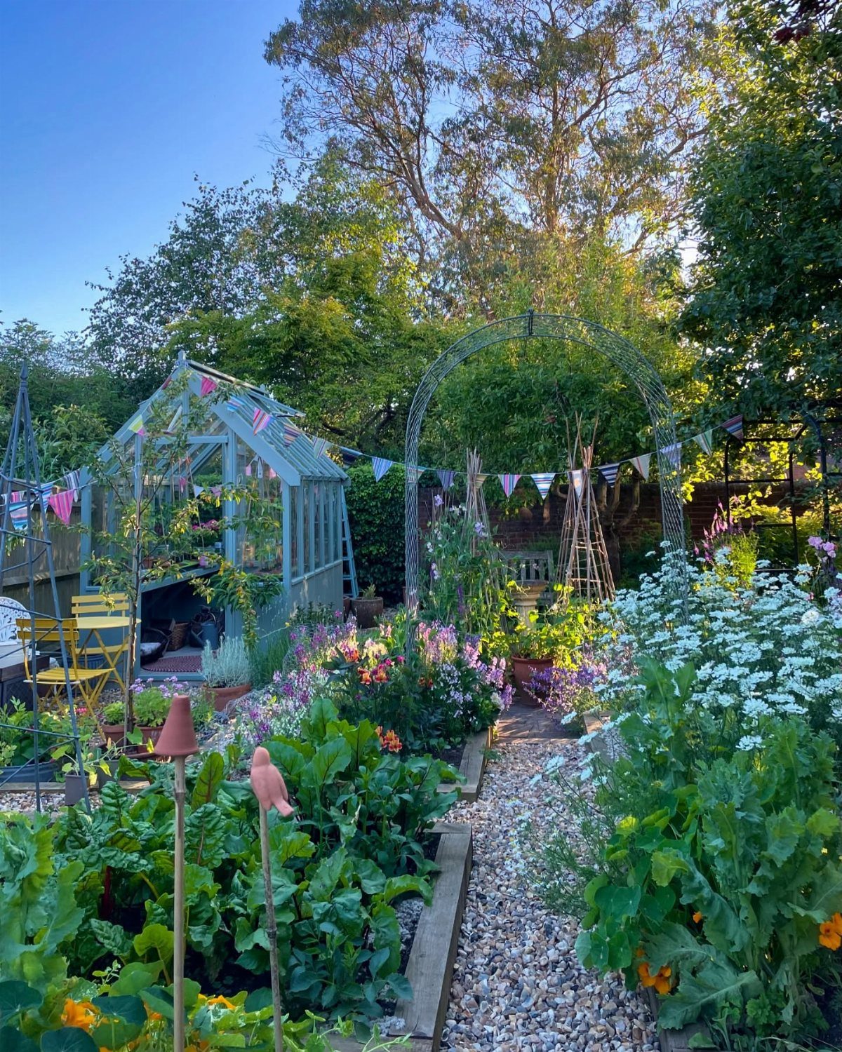 Vegetable garden with colorful flowers, a gravel path, and a blue greenhouse.