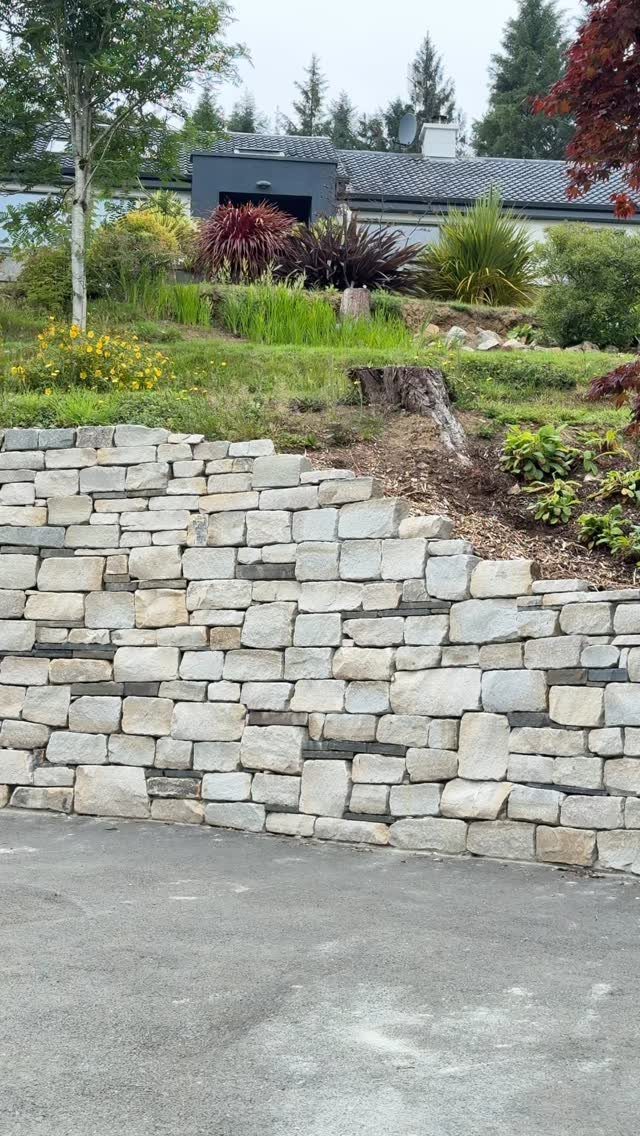 Stone retaining wall holding back a grassy slope with ornamental plants and shrubs.