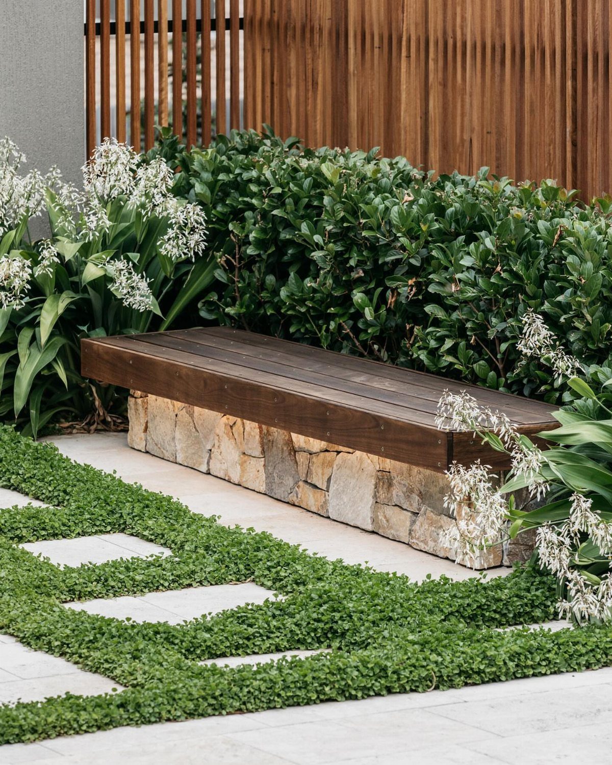 Wooden bench with stone base along a paved walkway with green ground-cover plants.
