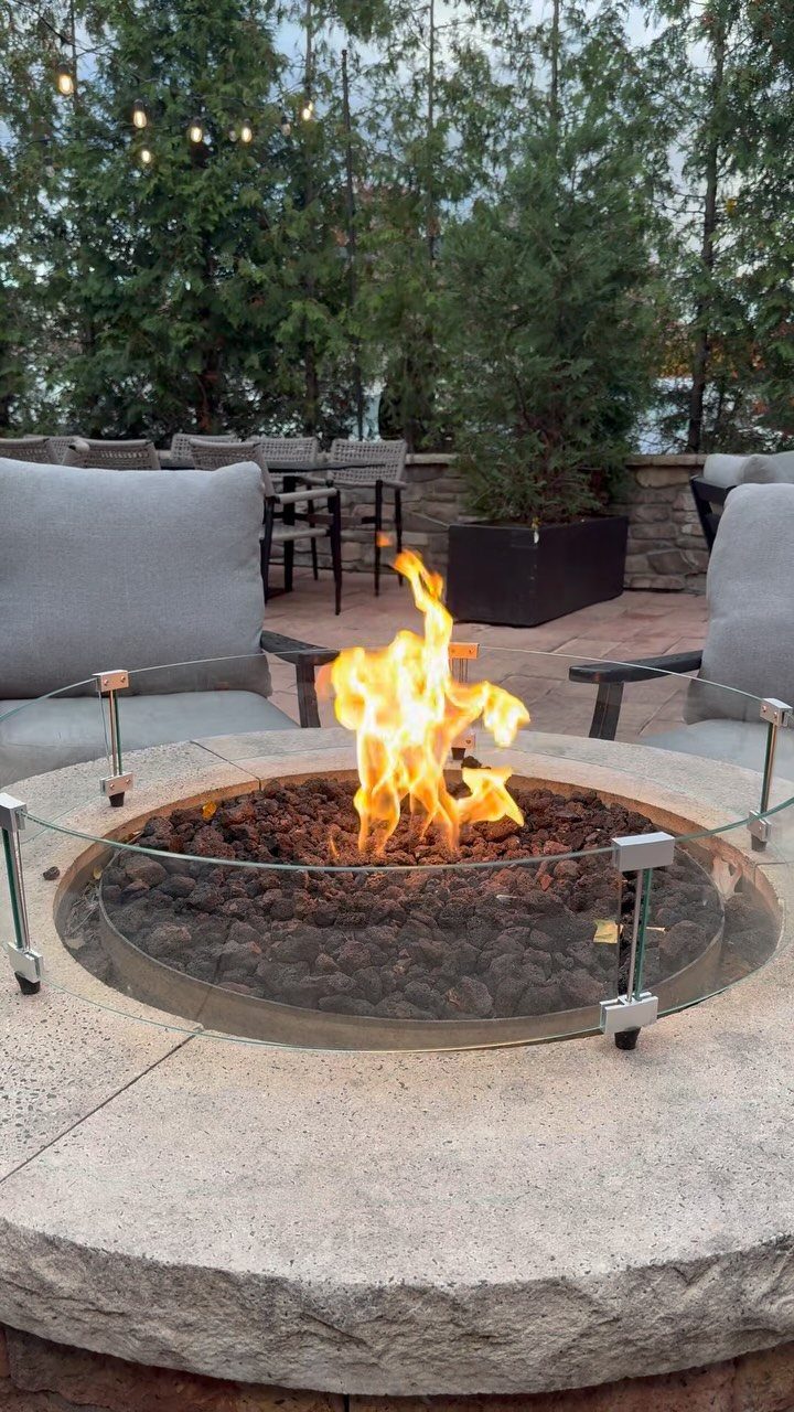 Circular stone fire pit with flames surrounded by grey outdoor armchairs on a patio.