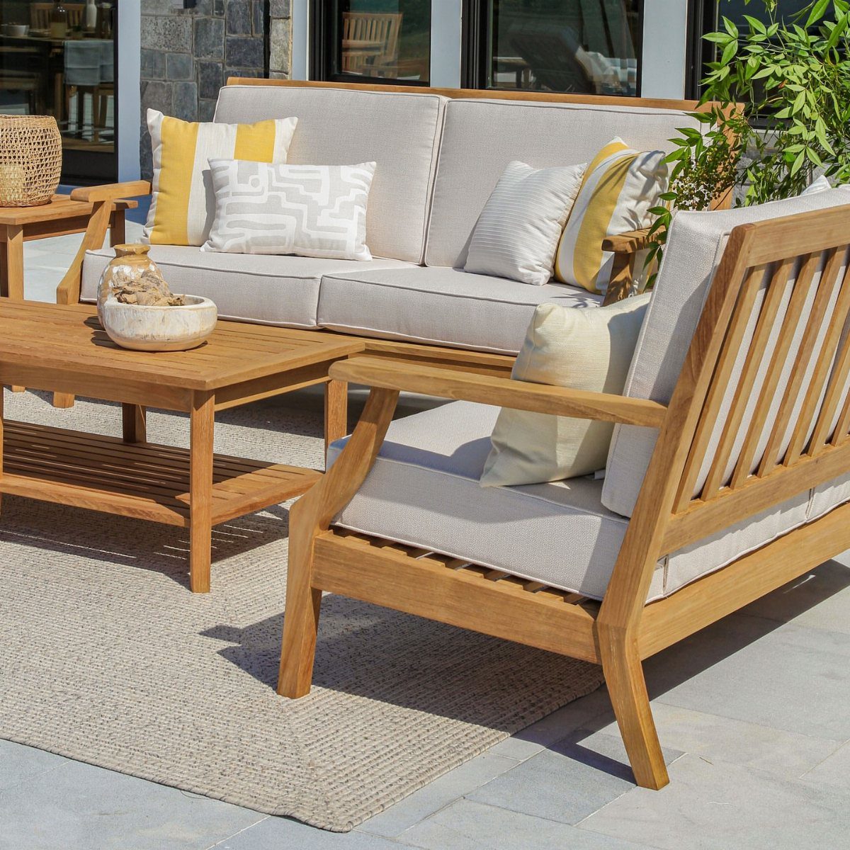 Long teak sofa and armchair with grey cushions and yellow accent pillows on a rug.
