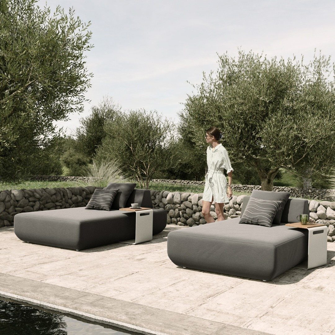 Two grey outdoor chaise longues with side tables on a stone patio by a pool.