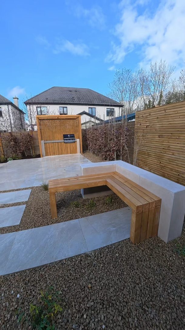 L-shaped wooden garden bench with a white stone backrest in a gravel courtyard.