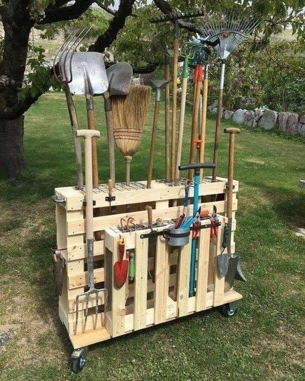Pallet wood garden tool organizer on wheels holding rakes and shovels outdoors.